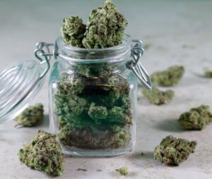 Glass jar filled with cannabis flower buds, with additional buds scattered on a white surface nearby.