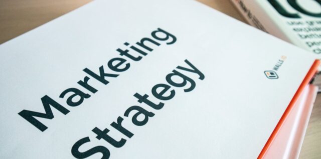 Marketing Strategy document cover with bold text, red spine binding, resting on wooden desk beside a book.