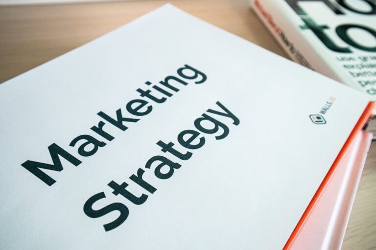 Marketing Strategy document cover with bold text, red spine binding, resting on wooden desk beside a book.