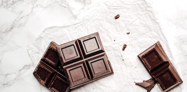 Broken dark chocolate bar pieces on white crinkled paper and marble surface with scattered crumbs